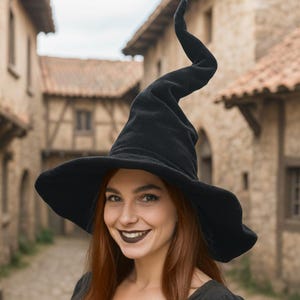 以下が含まれることがあります： 幅広のつばと高く湾曲した先端が付いた黒い魔女の帽子。帽子は柔らかく暗い生地で作られています。画像内の人物は笑顔で、濃い色の口紅を塗っています。背景は、古い建物のあるぼやけた通りのシーンです。