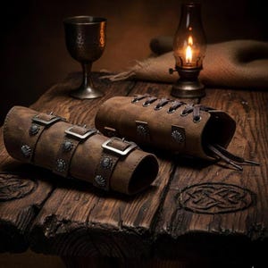 May include: Two brown leather arm guards with metal buckles and decorative studs. One guard is laced up, the other has three buckles. The guards are on a rustic wooden surface, suggesting a medieval or fantasy theme.