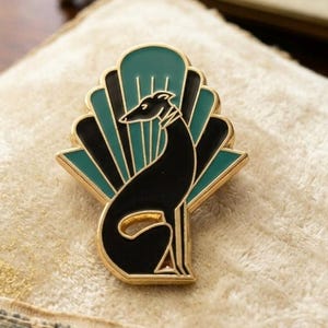 Puede incluir: Broche dorado con la silueta de un galgo negro. El broche tiene un fondo en forma de abanico en color verde azulado y negro. El perro está sentado con la cabeza alta. El broche es probablemente de metal y esmalte.