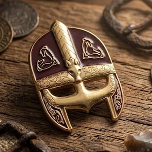 May include: A gold-toned metal pin shaped like a Viking helmet with a central ridge and a nose guard. The helmet features deep maroon enamel sections decorated with stylised animal motifs and intricate scrollwork patterns along the brow.