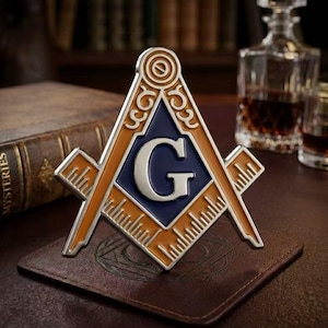 Könnte beinhalten: Ein dreieckiges Freimaurer-Emblem mit einem zentralen blauen Feld und einem silbernen Buchstaben G, umgeben von einem goldfarbenen Rand. Das Design zeigt Winkel und Zirkel mit sichtbaren Messmarkierungen entlang der Kanten der Werkzeugarme.