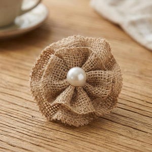 May include: A handcrafted burlap flower with a large, round, white pearl center. The flower has multiple layers of textured burlap petals. The flower is sitting on a wooden surface.