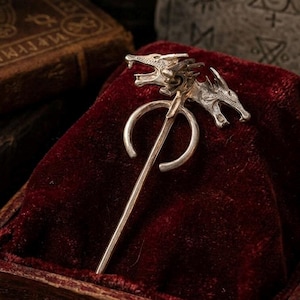 Puede incluir: Un broche de metal plateado que presenta dos cabezas de dragón orientadas en direcciones opuestas, unidas a un marco circular con un alfiler largo y recto que se extiende hacia abajo desde el centro del diseño.