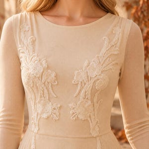 May include: A long-sleeved beige dress featuring a round neckline and intricate white floral embroidery on the bodice. The fabric has a textured, mesh-like appearance, and the floral appliqu&eacute;s extend from the shoulders down to the waistline.