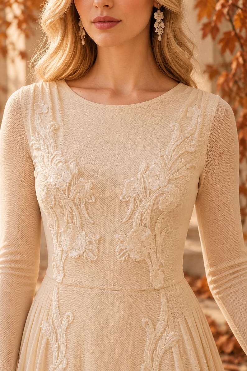 May include: A long-sleeved beige dress featuring a round neckline and intricate white floral embroidery on the bodice. The fabric has a textured, mesh-like appearance, and the floral appliqu&eacute;s extend from the shoulders down to the waistline.