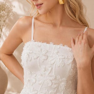 May include: A white sleeveless dress featuring a square neckline and thin spaghetti straps. The fabric is adorned with a textured, raised floral pattern with frayed edges throughout the bodice. The garment has a structured fit and clean lines.