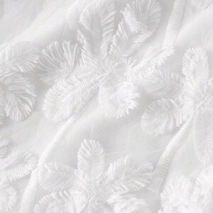 May include: A white fabric featuring a dense, raised floral embroidery pattern. The design consists of multiple flower motifs with textured, fringed petals and stems, all rendered in a monochromatic white thread on a matching white base.