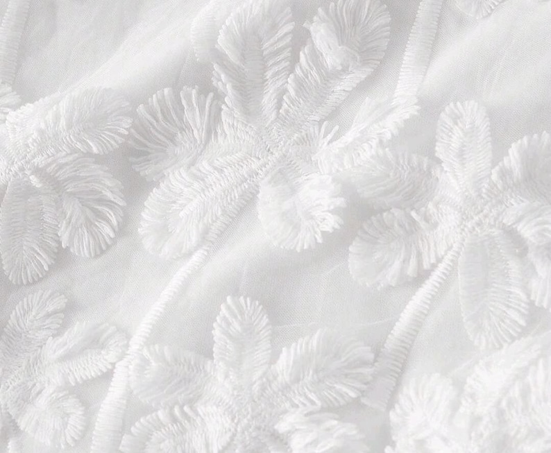 May include: A white fabric featuring a dense, raised floral embroidery pattern. The design consists of multiple flower motifs with textured, fringed petals and stems, all rendered in a monochromatic white thread on a matching white base.