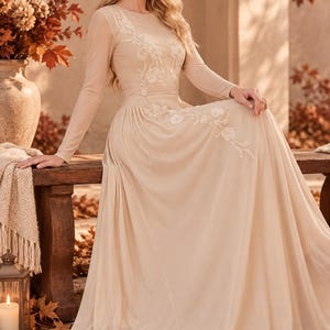 May include: A floor-length beige gown featuring long sleeves, a round neckline, and a sheer bodice overlay. The dress is adorned with delicate white floral embroidery that extends from the chest down to the waistline, flowing into a full skirt.