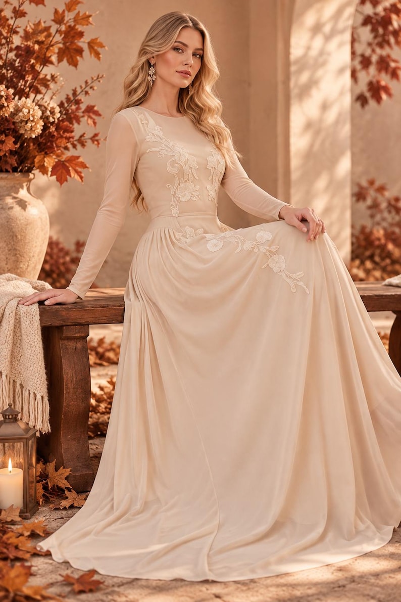 May include: A floor-length beige gown featuring long sleeves, a round neckline, and a sheer bodice overlay. The dress is adorned with delicate white floral embroidery that extends from the chest down to the waistline, flowing into a full skirt.