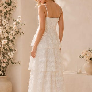 May include: A white midi-length dress featuring thin spaghetti straps, a fitted bodice, and a tiered skirt. The entire garment is covered in a delicate, raised floral appliqu&eacute; pattern, creating a textured appearance throughout the layered silhouette.