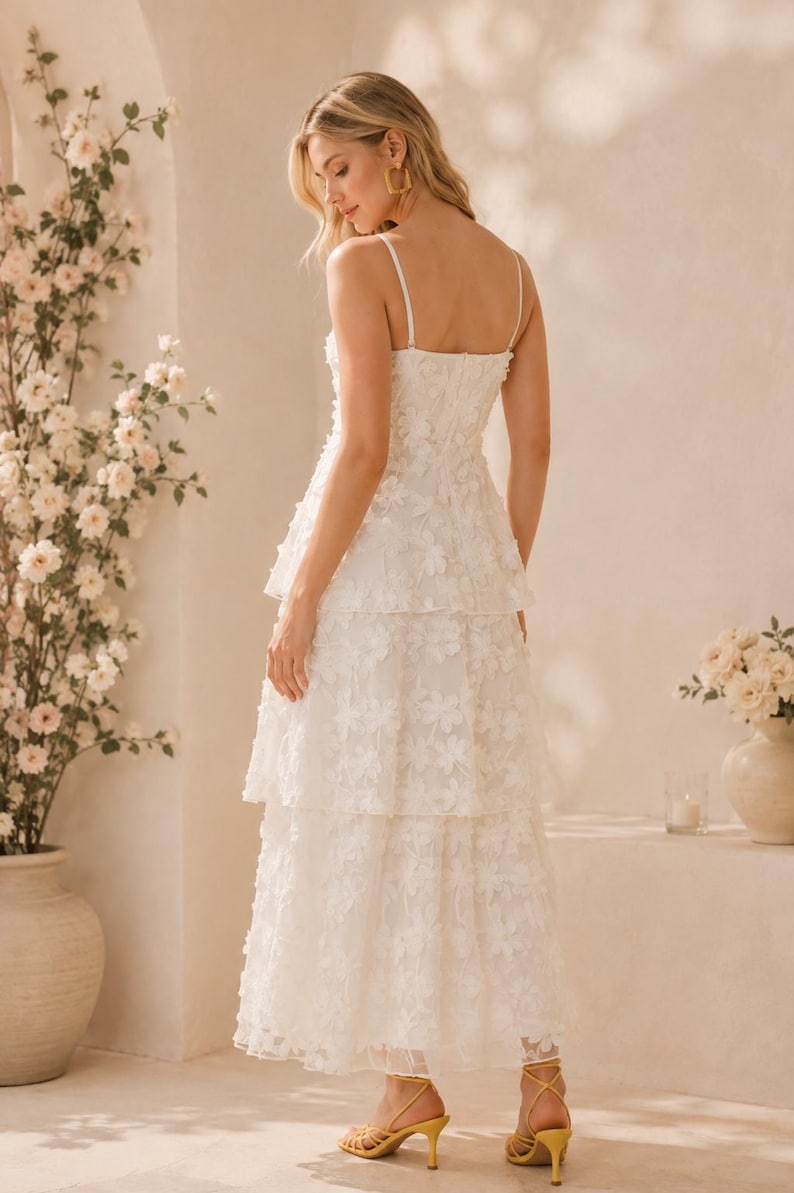 May include: A white midi-length dress featuring thin spaghetti straps, a fitted bodice, and a tiered skirt. The entire garment is covered in a delicate, raised floral appliqu&eacute; pattern, creating a textured appearance throughout the layered silhouette.