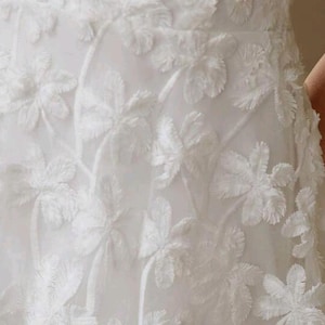 May include: A white garment featuring a textured, raised floral pattern across the fabric. The design consists of repeating flower motifs with fringed edges, creating a layered, three-dimensional appearance on the surface of the white material.