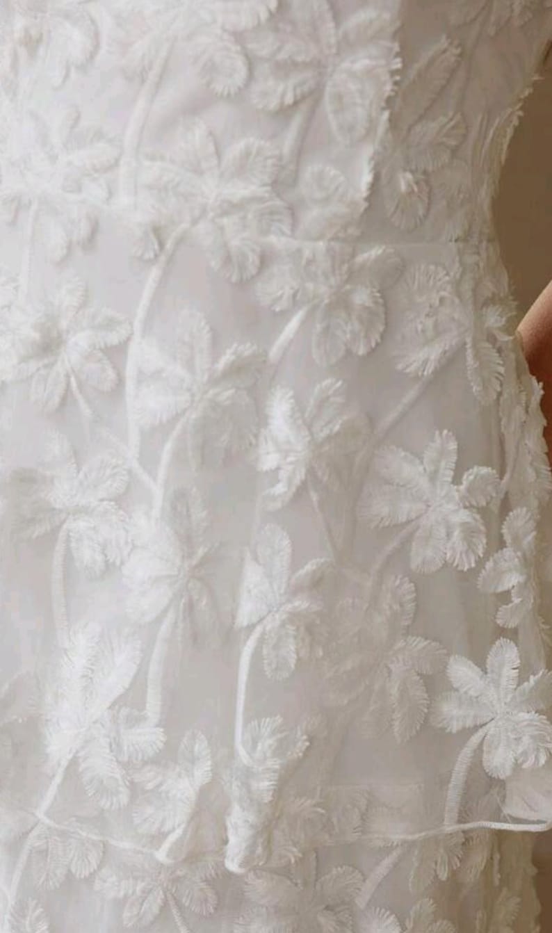 May include: A white garment featuring a textured, raised floral pattern across the fabric. The design consists of repeating flower motifs with fringed edges, creating a layered, three-dimensional appearance on the surface of the white material.