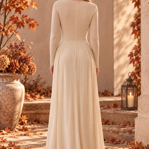 May include: A floor-length, long-sleeved dress in a solid beige color. The garment features a fitted bodice with a vertical center seam, a defined waistline, and a flowing, floor-length skirt. The back of the dress includes a visible vertical zipper.