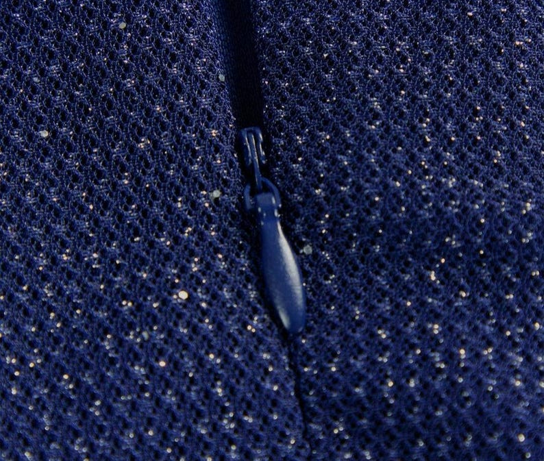 May include: A dark blue fabric with a fine mesh texture and subtle silver glitter particles throughout. A matching dark blue zipper pull is centered vertically, integrated into the seam of the material, showing a smooth, teardrop-shaped design.