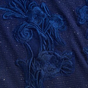 May include: A deep navy blue fabric featuring a dense, tonal floral embroidery pattern. The textile has a subtle, integrated shimmer throughout the weave, while the raised, stitched floral motifs create a textured, layered appearance across the surface.