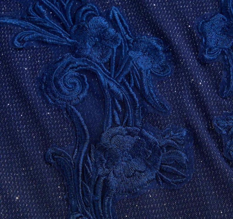 May include: A deep navy blue fabric featuring a dense, tonal floral embroidery pattern. The textile has a subtle, integrated shimmer throughout the weave, while the raised, stitched floral motifs create a textured, layered appearance across the surface.