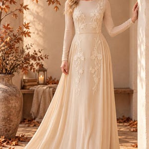 May include: A floor-length beige gown with long sleeves and a round neckline. The dress features sheer fabric on the upper bodice and sleeves, adorned with white floral embroidery that extends from the chest down to the waist and skirt area.