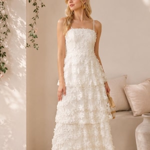 May include: A floor-length white dress featuring a square neckline, thin spaghetti straps, and a tiered skirt. The entire garment is covered in a delicate, three-dimensional floral appliqu&eacute; pattern, creating a textured and layered appearance.