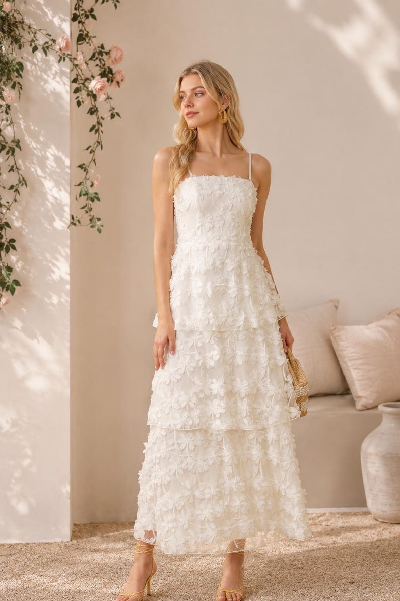 May include: A floor-length white dress featuring a square neckline, thin spaghetti straps, and a tiered skirt. The entire garment is covered in a delicate, three-dimensional floral appliqu&eacute; pattern, creating a textured and layered appearance.
