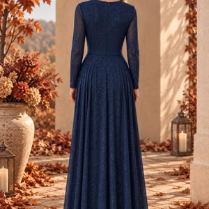 May include: A floor-length navy blue gown with long sleeves and a subtle glitter finish. The dress features a fitted bodice, a high neckline, and a pleated skirt that flows from the waist to the floor, creating a structured and elegant silhouette.