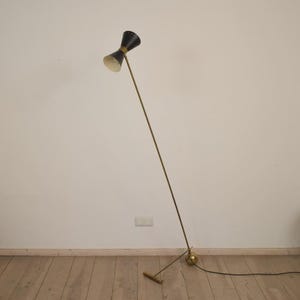 May include: A mid-century modern floor lamp with a black hourglass-shaped shade and a brass-colored stem. The lamp has a round base and a small rectangular foot. The lamp is plugged into an electrical outlet.