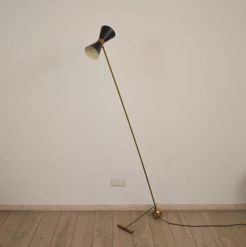 May include: A mid-century modern floor lamp with a black hourglass-shaped shade and a brass-colored stem. The lamp has a round base and a small rectangular foot. The lamp is plugged into an electrical outlet.