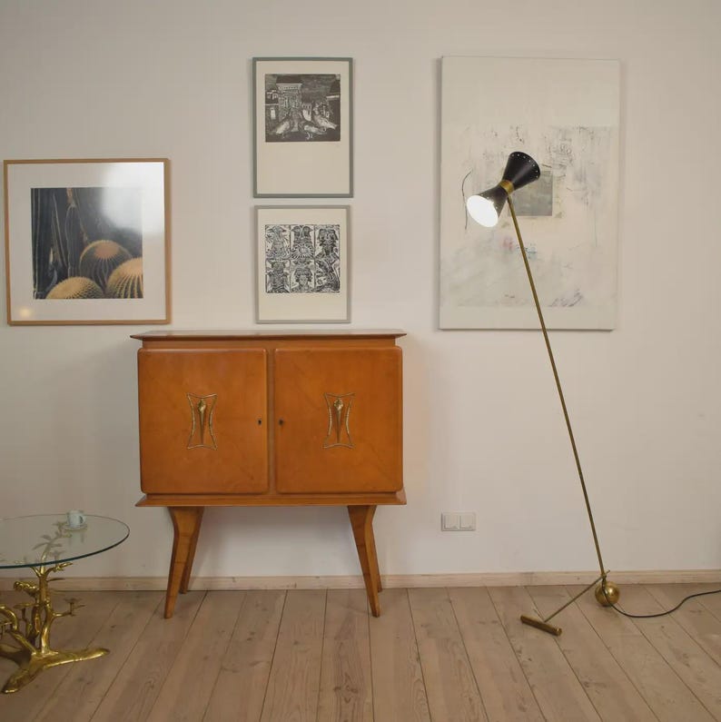 May include: A mid-century modern interior featuring a wooden cabinet with decorative gold accents, framed artwork, and a black and gold floor lamp. A small glass table with a gold base is in the foreground. The room has hardwood floors and white walls.