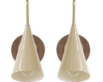 May include: A pair of cream-coloured, cone-shaped wall sconces with brass accents and a wooden backplate. The sconces have a mid-century modern design. The light bulbs are visible inside the sconces. The design is simple and elegant.