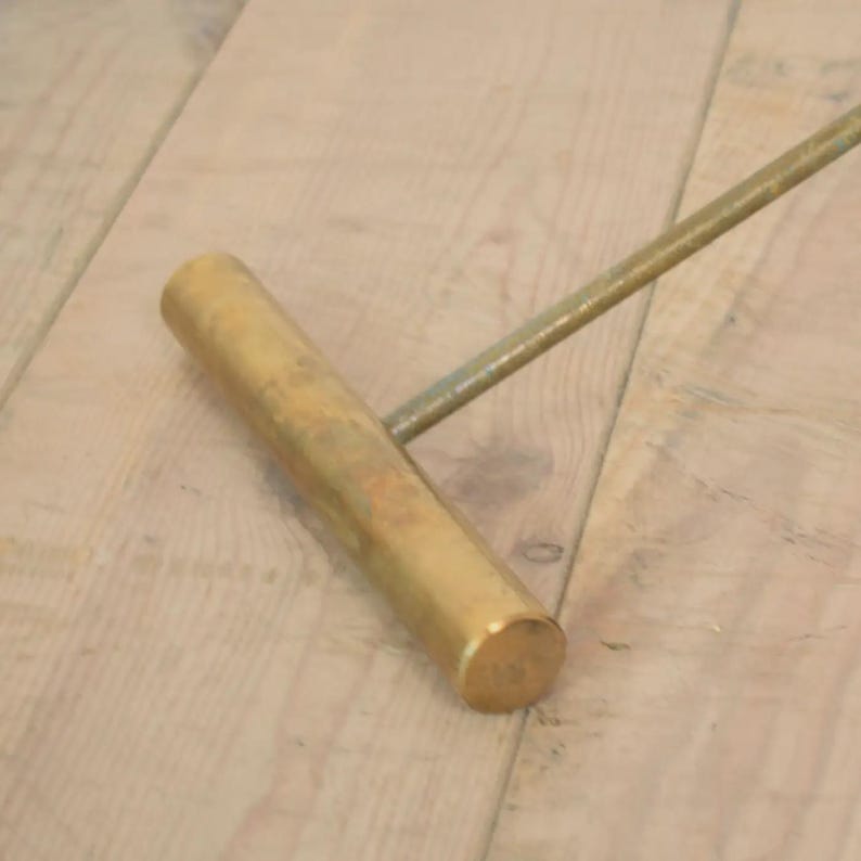 May include: A brass-colored, cylindrical handle with a connecting rod. The handle is positioned diagonally across a light-colored wooden surface. The rod extends from the handle at an angle, suggesting a tool or hardware component.