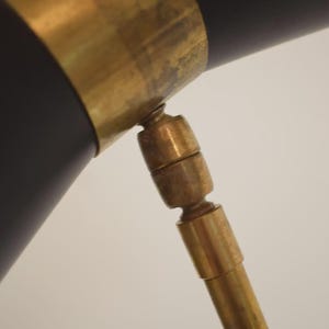 May include: Close-up of a black and brass desk lamp. The lamp features a black cylindrical shade with a brass band and a brass adjustable arm. The design is a mid-century modern style.