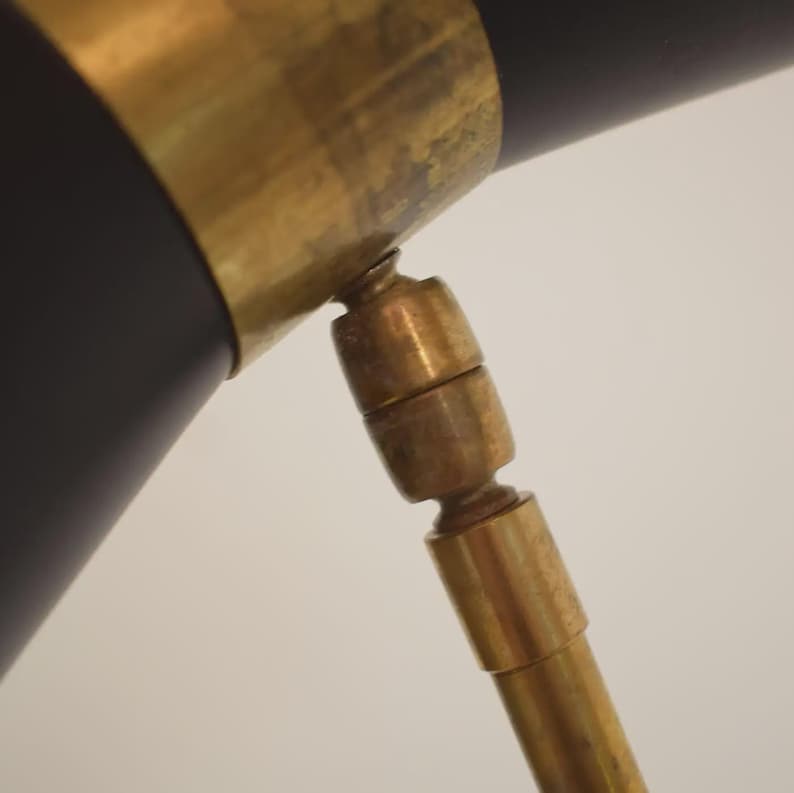 May include: Close-up of a black and brass desk lamp. The lamp features a black cylindrical shade with a brass band and a brass adjustable arm. The design is a mid-century modern style.