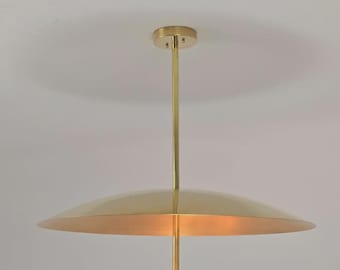 May include: A modern brass pendant light fixture. The light has a circular canopy, a long central rod, a large, inverted saucer-shaped shade, and a smaller, upward-facing bowl-shaped element below. The fixture is suspended from the ceiling.