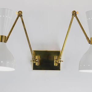 Può includere: Applique da parete bianco e oro con due lampade a forma di cono. Le lampade hanno una serie di piccoli fori vicino alla parte superiore e sono collegate da bracci dorati regolabili a una base rettangolare dorata.
