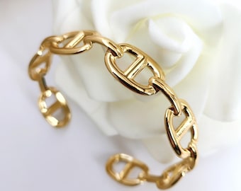 May include: A gold-colored chain link bracelet with a distinctive anchor-shaped design. The bracelet is displayed on a white surface with a white flower in the background. The bracelet is made of a shiny, reflective metal.