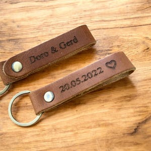 Könnte beinhalten: Zwei braune Leder-Schlüsselanhänger mit silbernen Schlüsselringen. Einer ist mit "Doro & Gerd" graviert, der andere mit "20.05.2022" und einem Herzsymbol. Die Schlüsselanhänger sind mit Metallnieten befestigt.