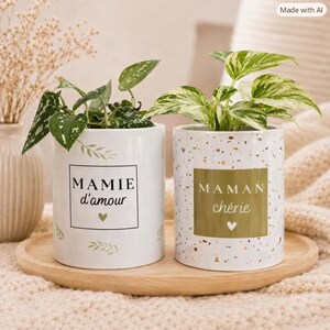 Peut inclure: Deux pots de fleurs cylindriques en céramique sur un plateau en bois. Le pot de gauche est blanc avec des motifs de feuilles vertes et le texte "MAMIE d'amour" avec un cœur. Le pot de droite est moucheté, avec un carré doré et "MAMAN chérie".