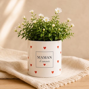 Könnte beinhalten: Ein weißer Keramik-Blumentopf mit roten Herzakzenten und dem Text "MAMAN d'amour" in einem schwarzen Quadrat. Der Topf ist mit grünem Laub und weißen Blüten gefüllt und steht auf einem beigen Tuch.