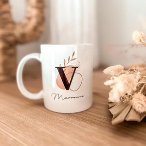 Peut inclure: Mug en céramique blanche avec la lettre noire "V" et le mot "Marraine" en écriture cursive. Le mug présente un motif avec des formes abstraites et des éléments botaniques dans des tons de marron et de pêche. La tasse a une anse incurvée.