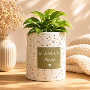 Op de afbeelding: Een witte keramische plantenpot met een gespikkeld ontwerp, met een groene plant. De pot heeft een rechthoekig label met de woorden "MAMAN chérie" en een hartsymbool. De pot staat op een houten oppervlak.