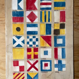 Op de afbeelding: Een rechthoekig bord met een handgeschilderd raster van zesentwintig maritieme signaalvlaggen, elk gelabeld met de bijbehorende fonetische alfabetnaam van Alpha tot Zulu. De vlaggen tonen diverse geometrische patronen in rood, blauw, geel en wit.