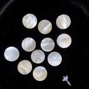 May include: A collection of ten circular mother-of-pearl buttons with a smooth, iridescent finish. Each button displays a unique, swirling white and cream pattern with a subtle sheen. A small, light purple flower is positioned near the bottom right.