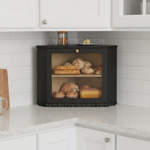 May include: A black corner bread box with a glass door, displaying various loaves of bread. The wooden interior has two shelves. A small jar and a plate with food are on top of the bread box. The bread box is on a white countertop.