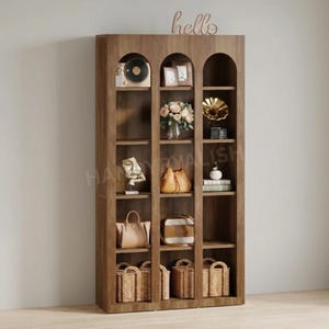 May include: A tall, wooden bookshelf with arched top shelves. The shelves display a variety of decorative items, including a vintage record player, framed photos, flowers, handbags, sculptures, and woven baskets. A cursive "hello" sign is mounted on top.