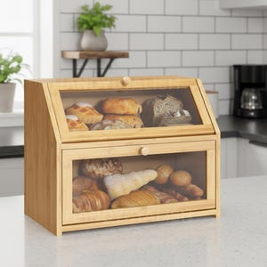 May include: A two-tiered bamboo bread box with clear glass doors. The top section has a slanted door, and the bottom has a pull-out door. The box is filled with various types of bread and pastries. The bread box is on a white countertop.