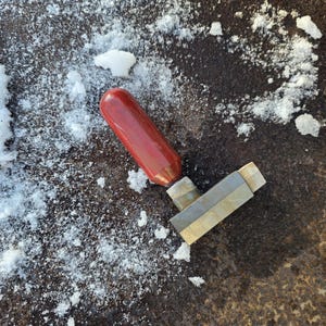 May include: A red-handled tool with a metallic, geometric head rests on a textured, dark surface, possibly stone or metal. Patches of white snow surround the tool, creating a contrast of color and texture. The tool appears to be a small chisel or similar implement.