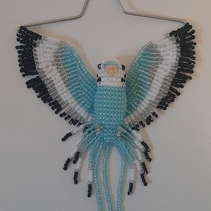 May include: A beaded bird ornament with outstretched wings, featuring a light blue body, white, silver, and black accents. The bird is suspended by a silver wire hanger, creating a decorative piece.