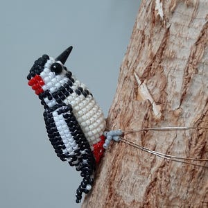 Peut inclure: Figurine de pic en perles noires, blanches et rouges, perchée sur un tronc d'arbre. L'oiseau a un bec et un œil noirs, avec une tache rouge sur la tête. Le pic est fixé à l'arbre avec du fil de fer.
