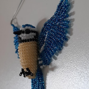 Pode incluir: Um enfeite de pega-azul de contas com as asas abertas. O pássaro apresenta um corpo bege, detalhes em preto e branco e uma cabeça e asas azuis. Um fio de prata é preso para pendurar.
